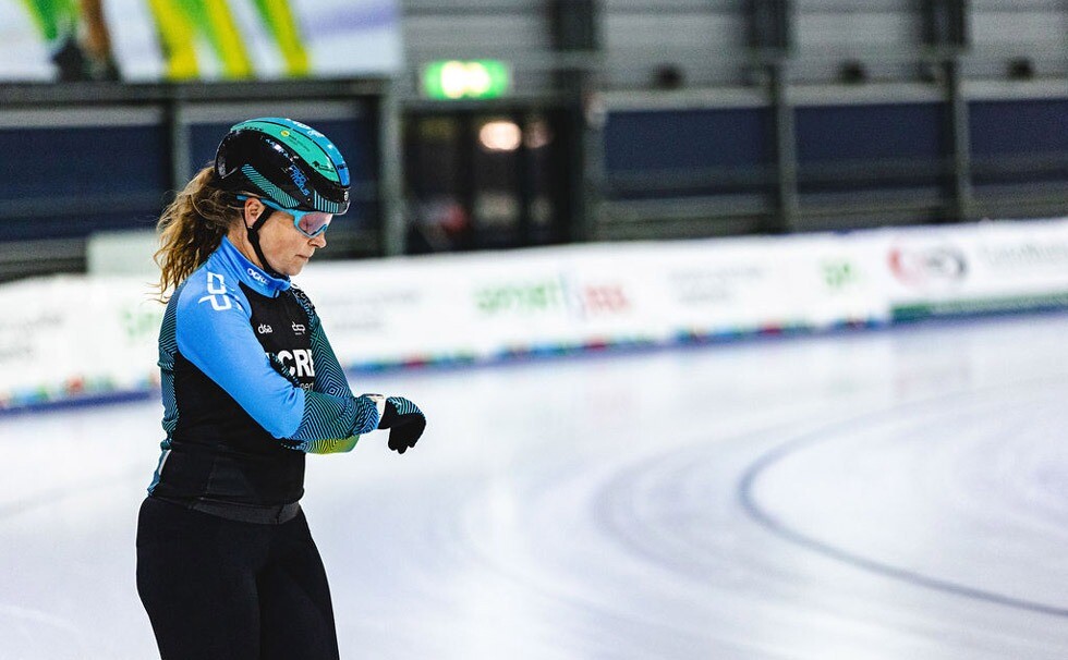Coach DCP/OCRE schaatsteam controleert tijd op ijsbaan Coach van het DCP/OCRE marathonschaatsteam staat op het ijs en kijkt geconcentreerd op haar horloge, gekleed in teamkleding met helm en bril.