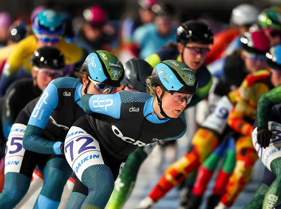 DCP/OCRE marathonschaatsteam tijdens massastart met rijdster 72 in actie Schaatsster van het DCP- en OCRE-gesponsorde team met nummer 72 in actie tijdens een drukbezette marathonwedstrijd, omringd door rijders in diverse pakken.