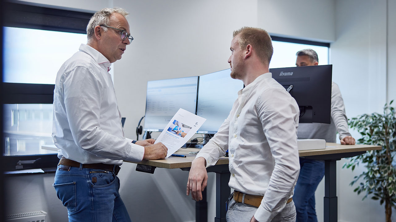 Lars Kiewiet van DCP in gesprek met een collega in de werkplaats, omringd door printers en apparatuur.